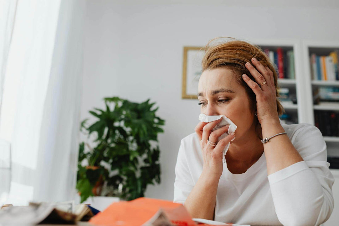 Frau mit Allergie putzt sich die Nase in ihrer Wohnung, im Hintergrund steht eine Pflanze.